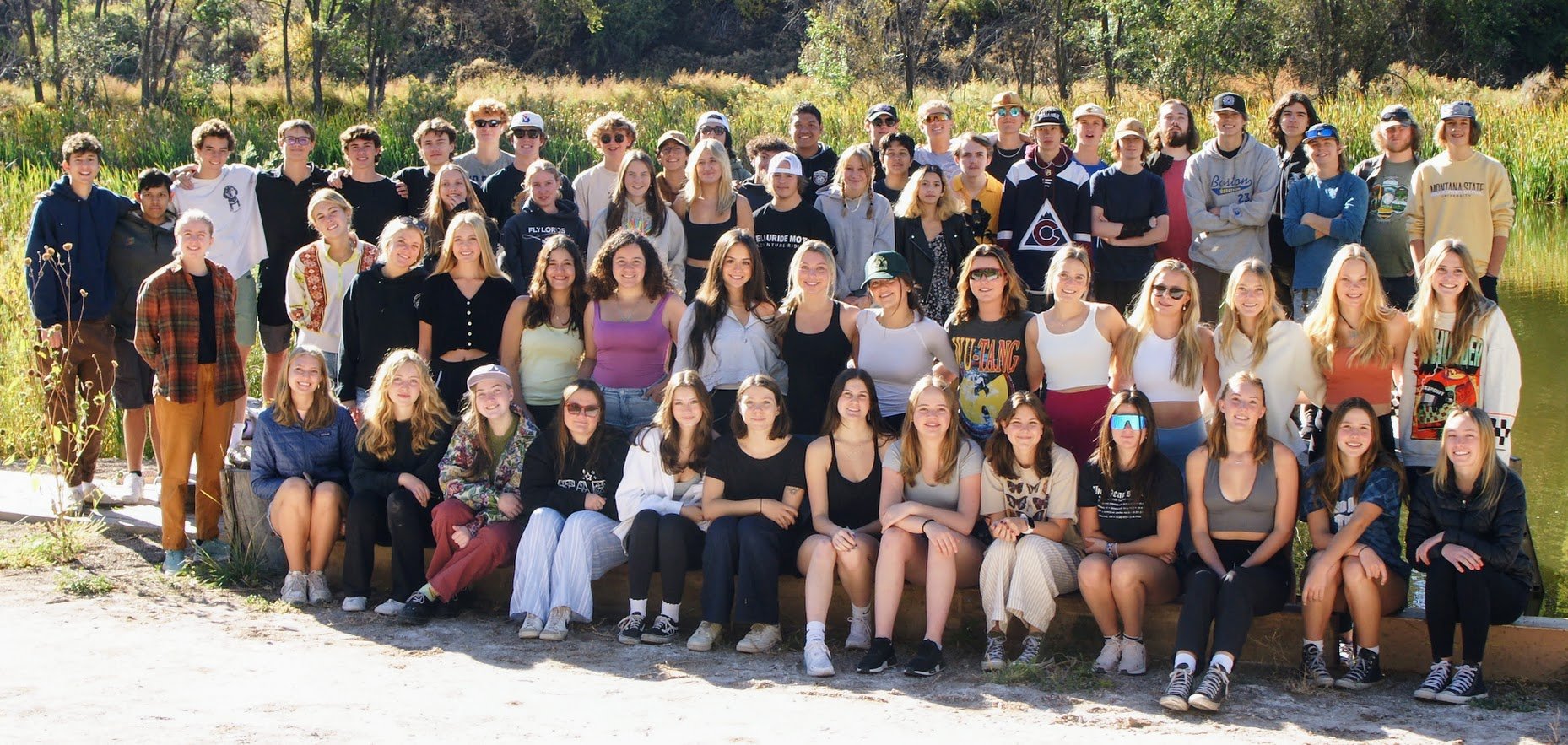 A group of people, mostly young adults, pose outdoors in front of trees and vegetation. They are dressed casually, some wearing hats and sunglasses. The setting appears to be a natural area, suggesting a sunny day.