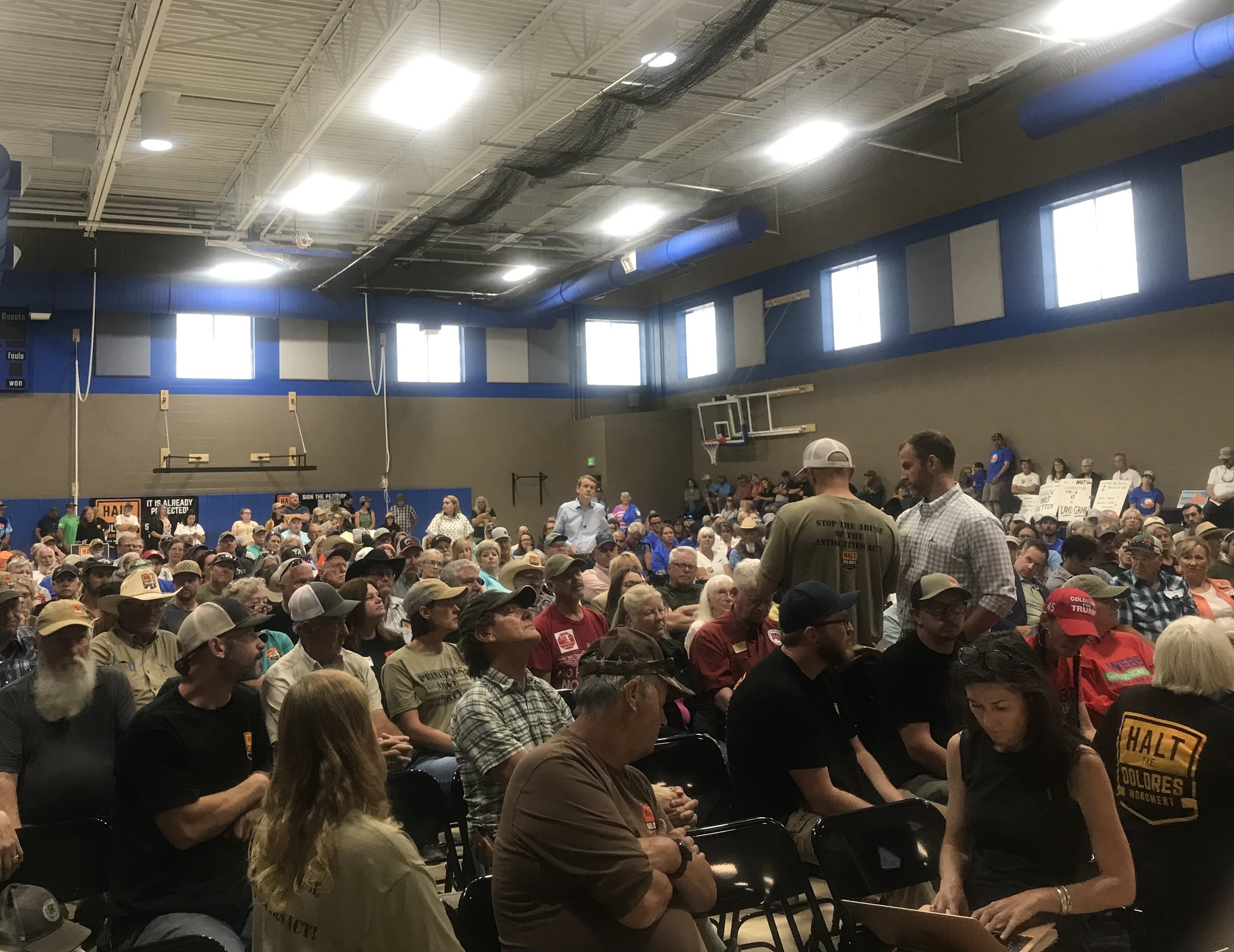 A large group of people seated in a gymnasium, many wearing hats. Some individuals are standing and conversing. The room is brightly lit with banners on the walls and a basketball hoop visible. The atmosphere appears to be an organized event or meeting.