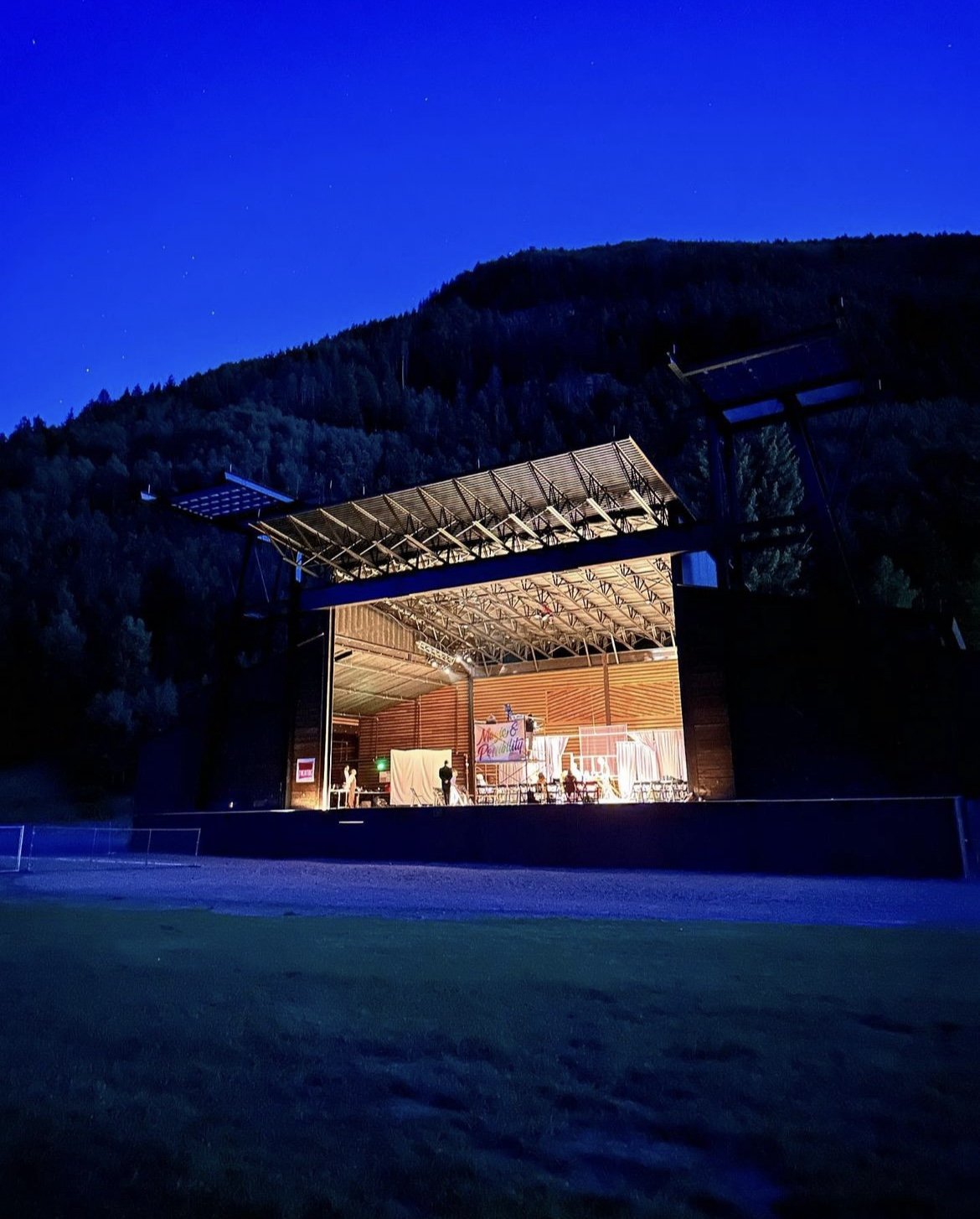 Outdoor stage illuminated against a dark mountain backdrop during dusk. Some people are visible on stage, with equipment and set pieces around them. The sky is deep blue.