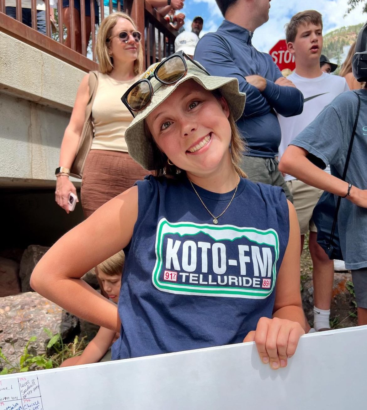 A smiling person wearing a bucket hat, sunglasses, and a KOTO-FM Telluride shirt stands outdoors holding a sign. Several people, including a woman with sunglasses and a phone, are gathered in the background near a wooden railing.