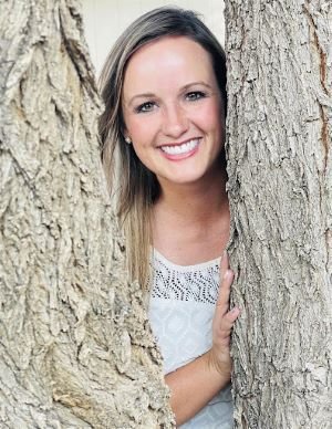 Smiling woman with long hair, wearing a white top, peeks through the gap between two tree trunks.