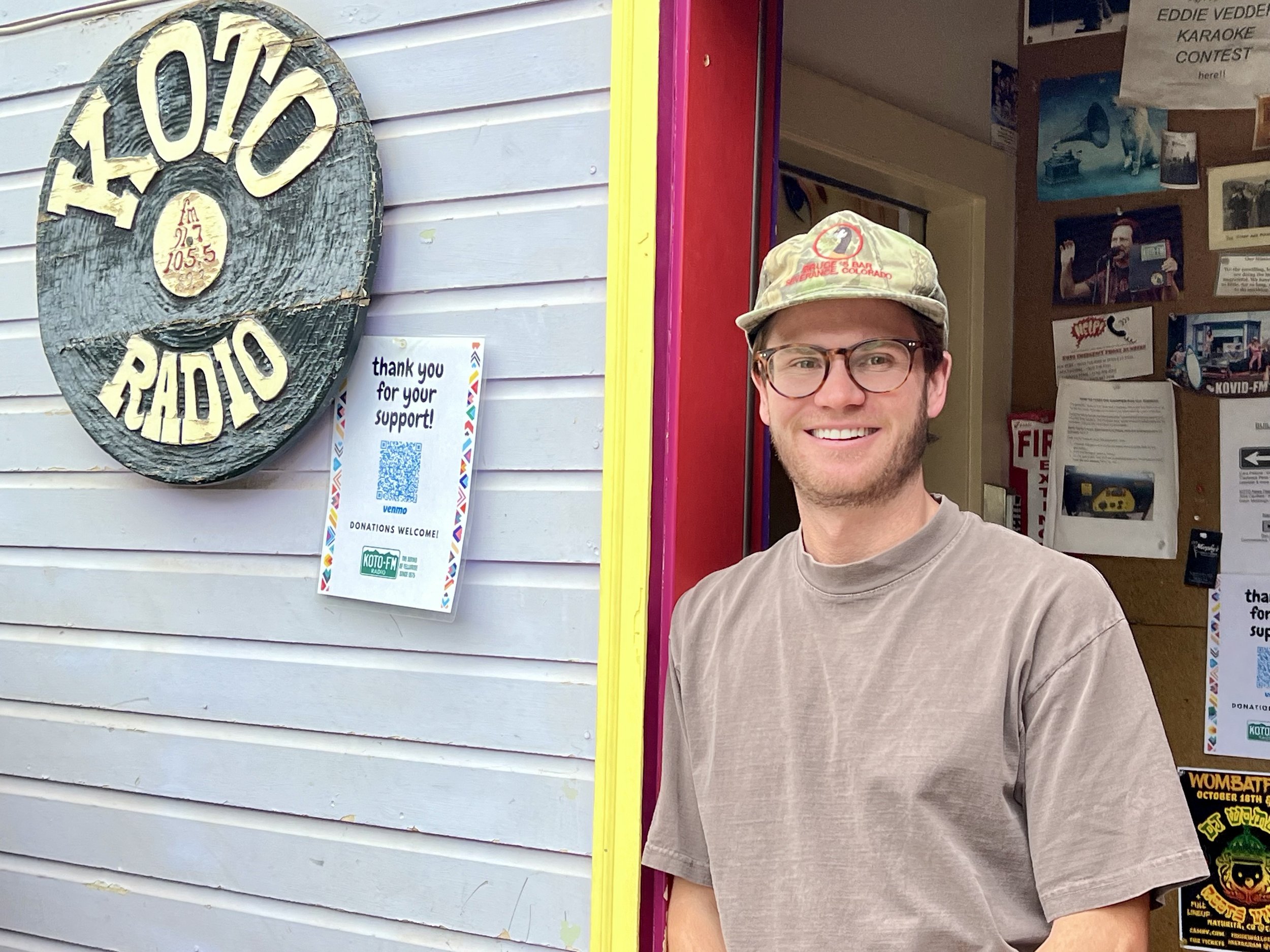 A person wearing glasses, a cap, and a brown T-shirt stands smiling in the doorway of a building. Next to them is a sign reading KOTO Radio above a vinyl record design. Posters and notes are visible on the wall inside.