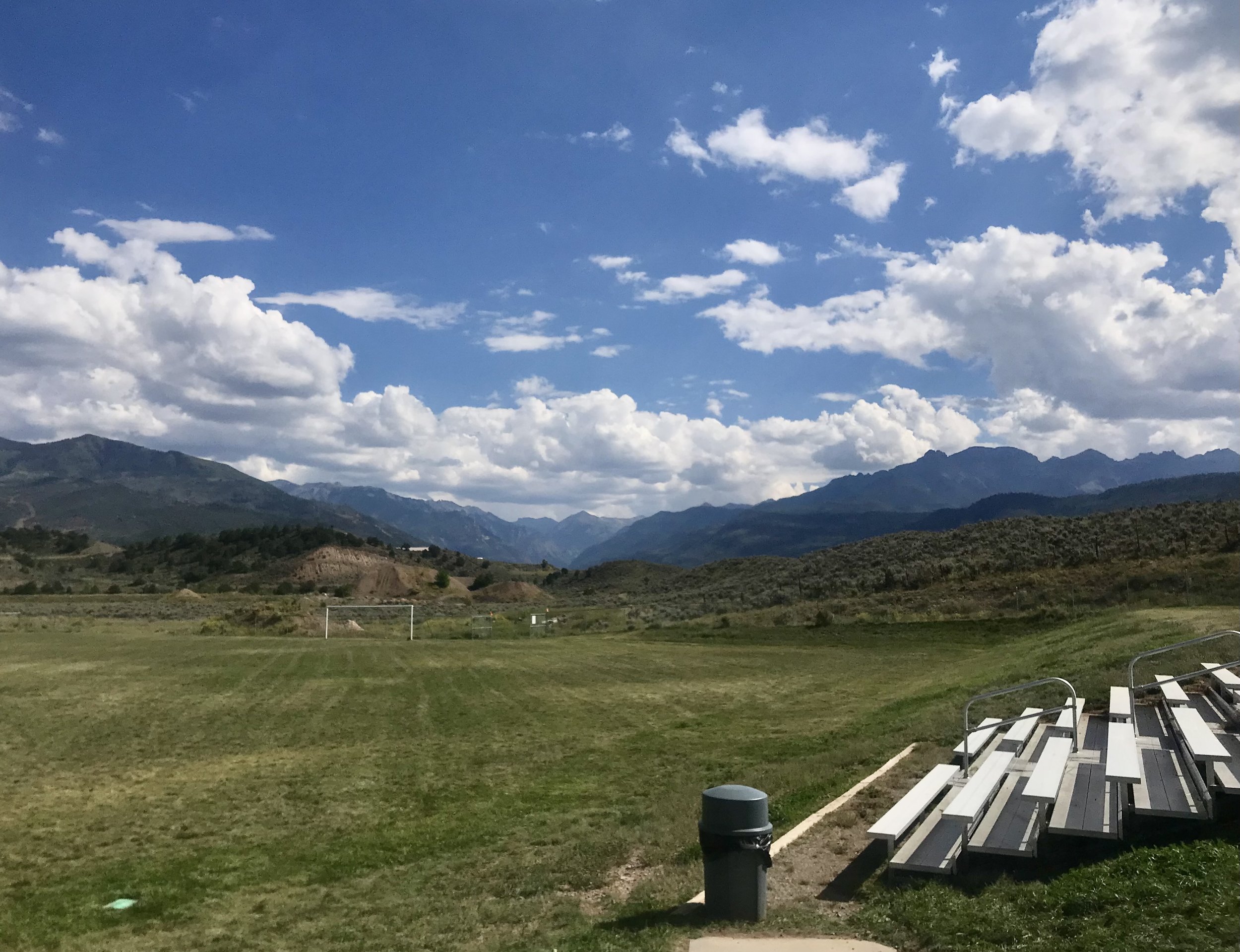 Bleachers overlook a grassy sports field under a blue sky with scattered clouds. Mountains rise in the distance, creating a scenic backdrop.