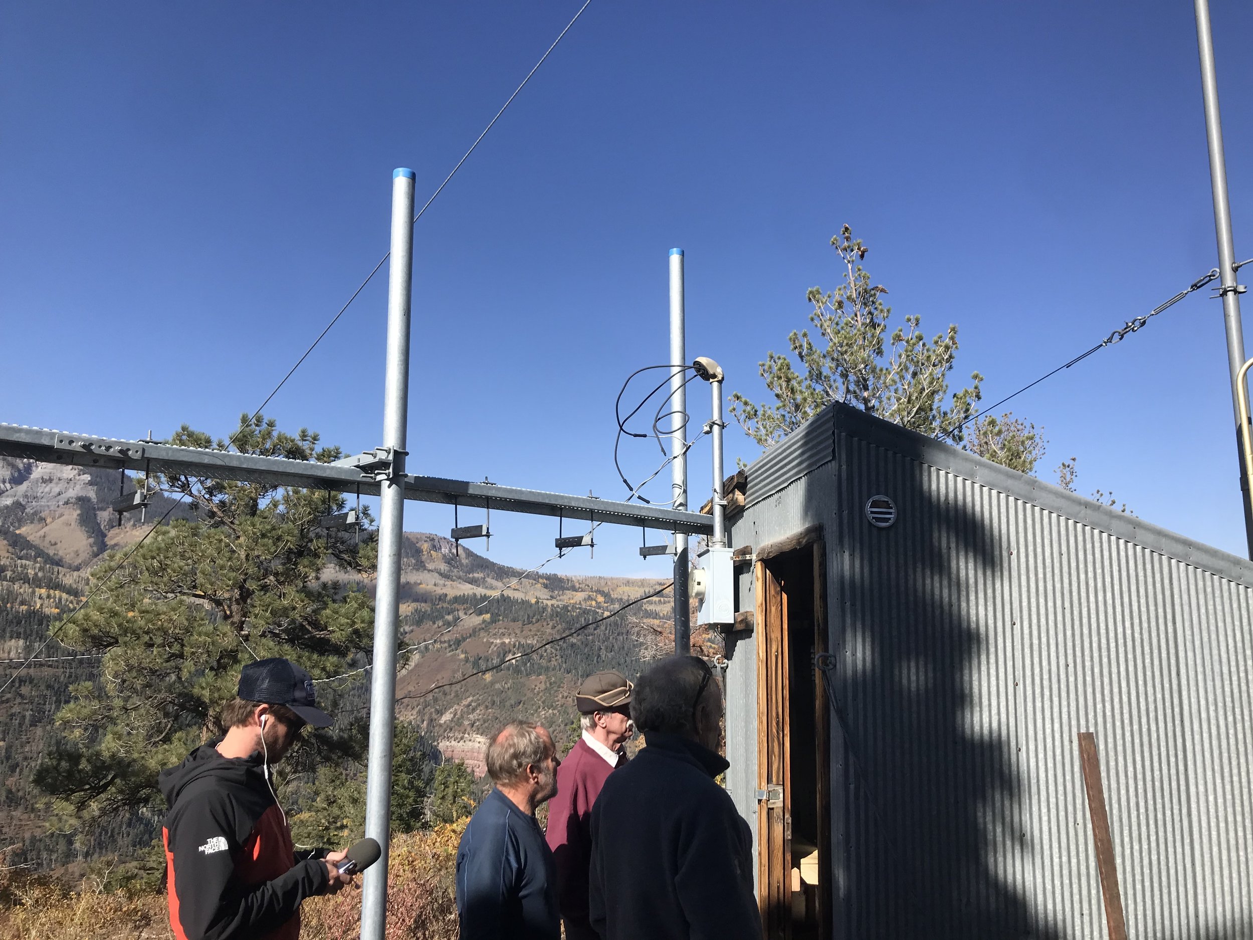 Four people stand outside a small corrugated metal building at a mountaintop facility surrounded by tall poles and wires. The sky is clear and blue, and mountains and trees are visible in the background.