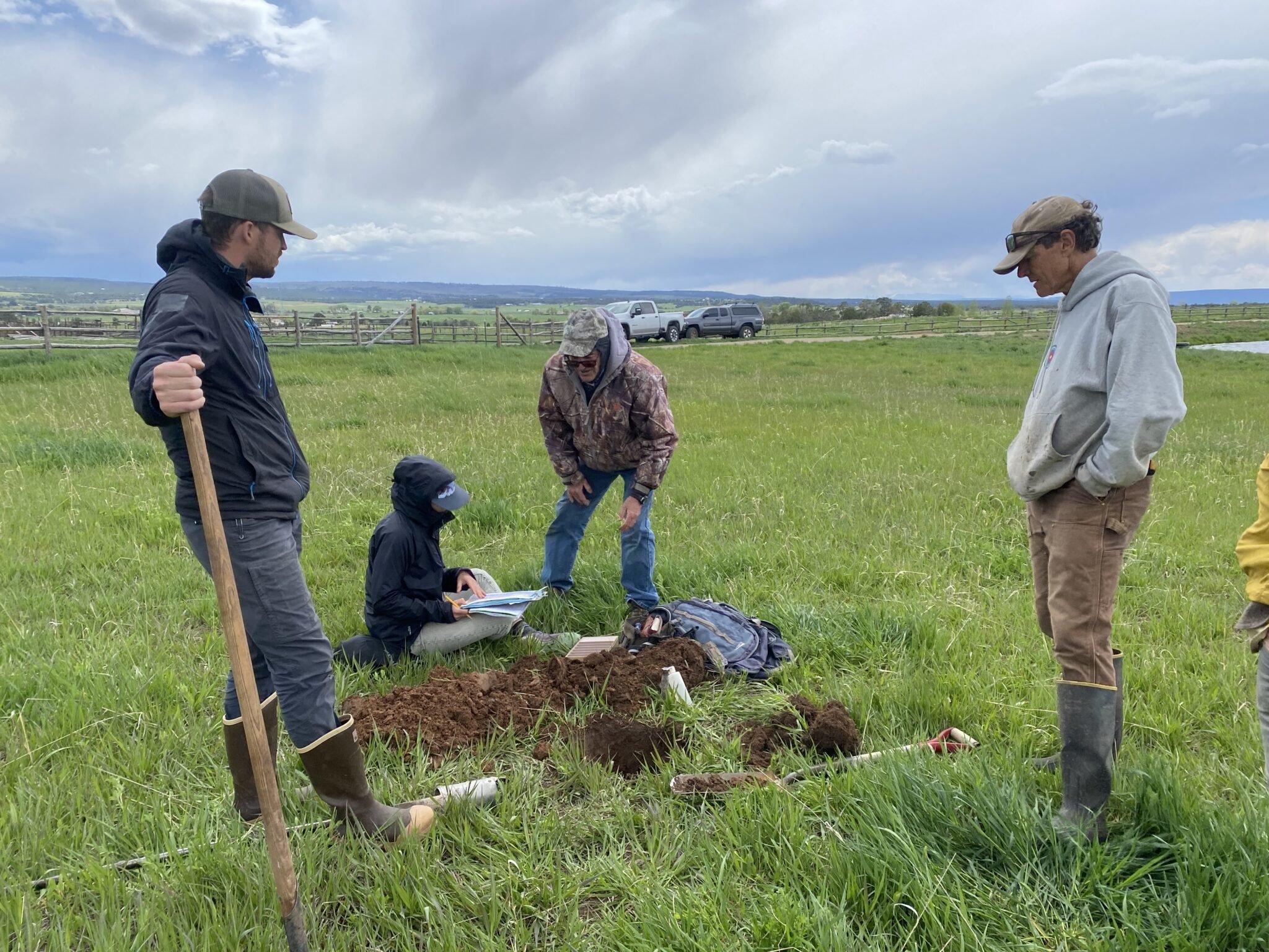 Cuatro personas examinan la tierra de un hoyo excavado en un campo cubierto de hierba. Uno sostiene una pala, otro escribe en un cuaderno, otros dos observan de pie. El cielo está nublado y hay colinas al fondo. Cerca se ven un camión y una valla.