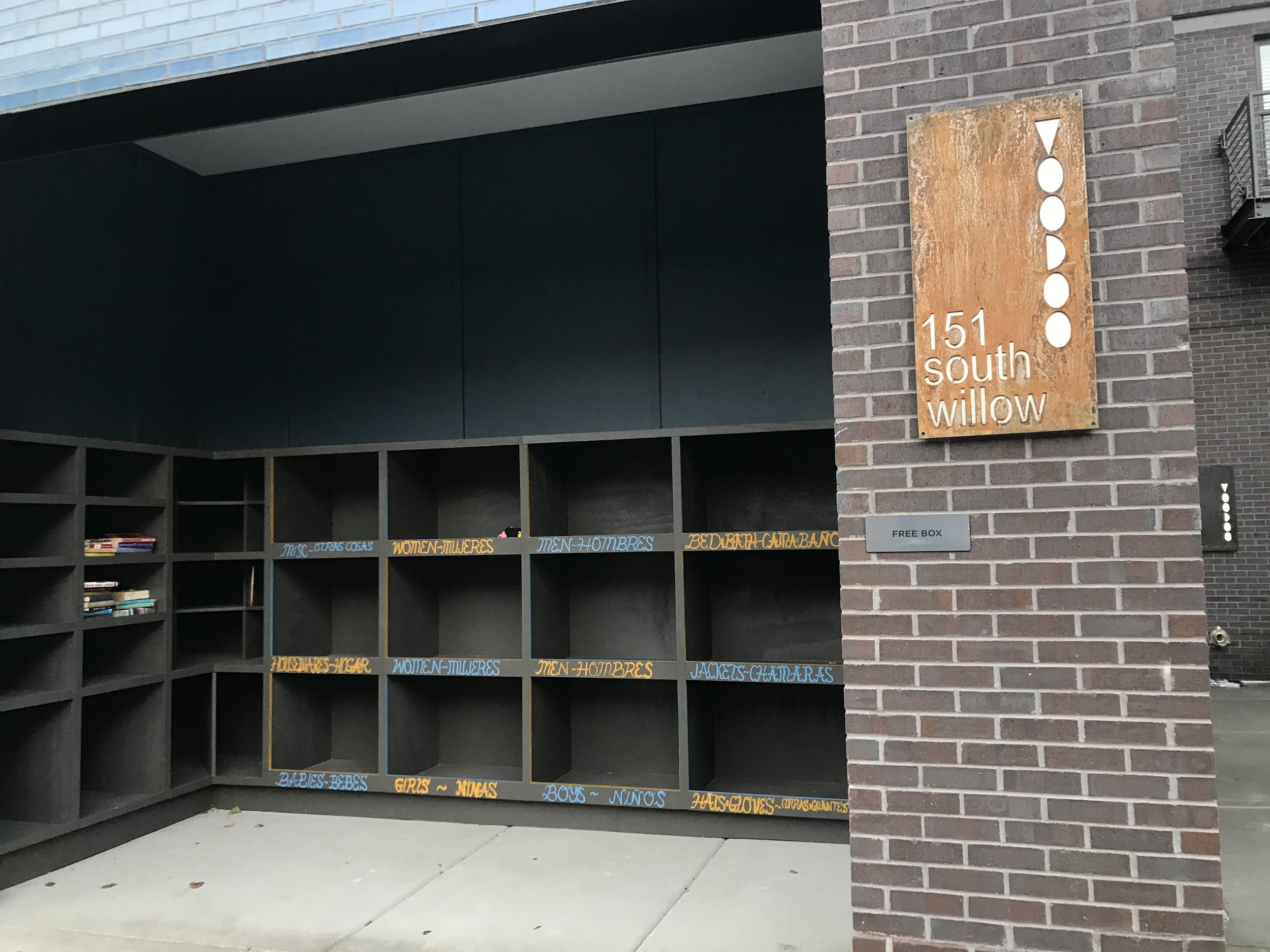 Una biblioteca minimalista al aire libre con estanterías oscuras que presentan una pequeña selección de libros. Situada junto a una pared de ladrillo con un letrero de metal oxidado que dice 151 South Willow. Un cartel más pequeño debajo dice Free Box.