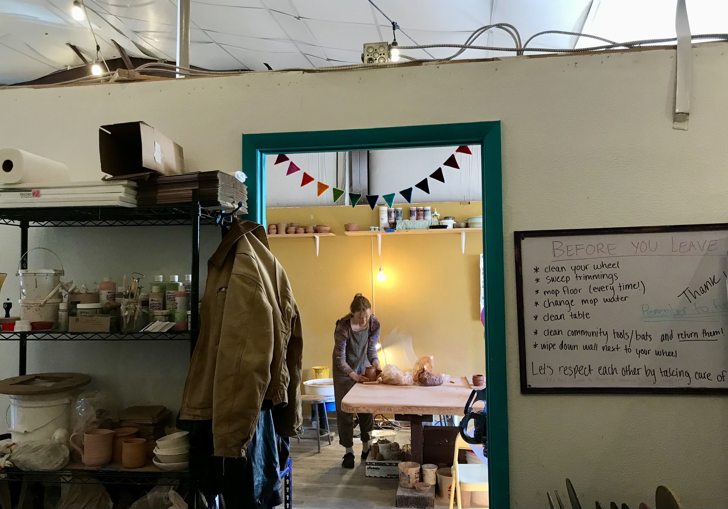 A person working at a pottery wheel in a room with yellow walls, visible through an open doorway. Shelves with supplies line the walls, and a chalkboard sign with instructions hangs nearby. Colorful flags are strung across the doorway.