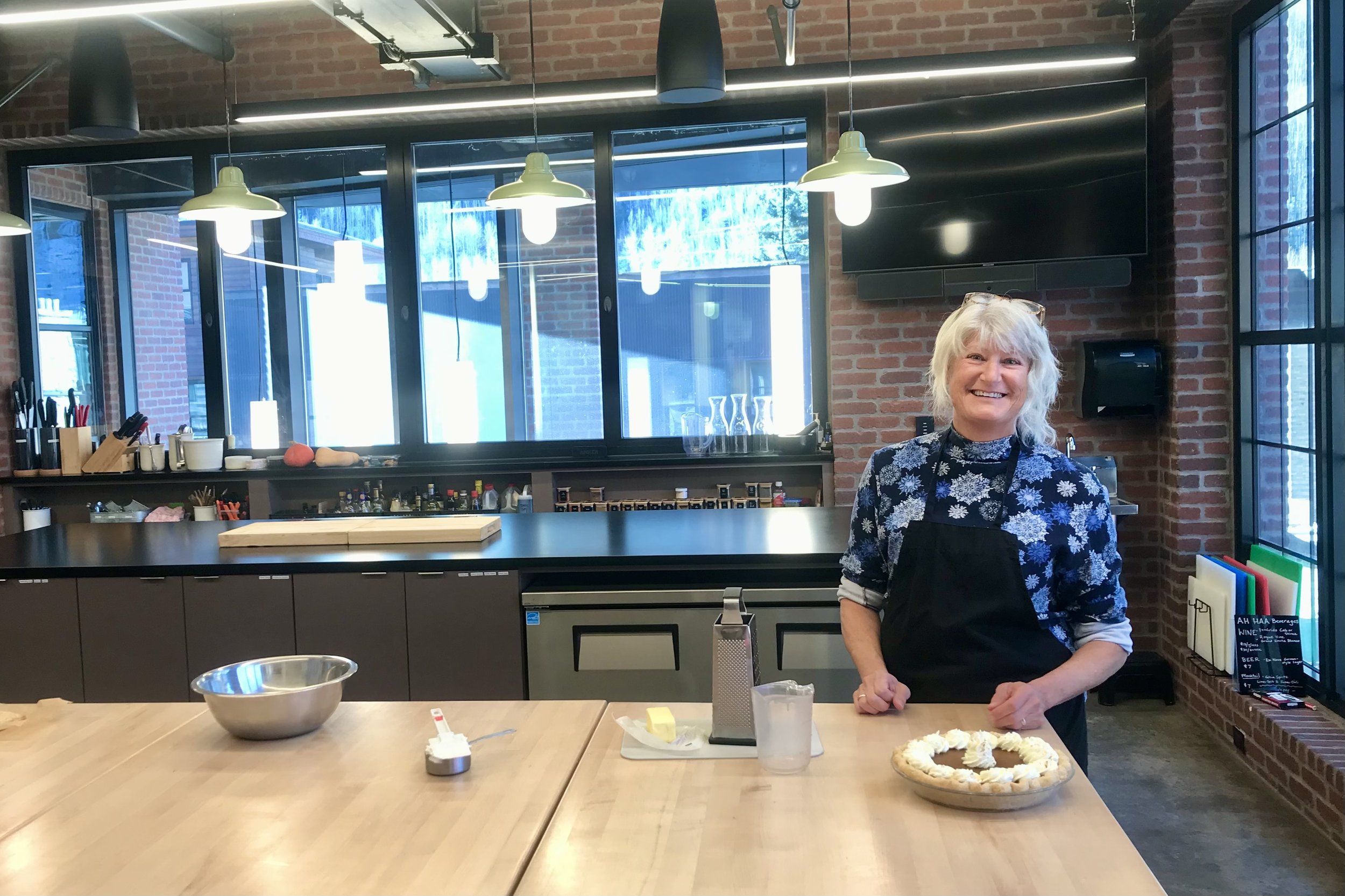 A woman with gray hair, wearing a black apron and floral shirt, stands in a modern kitchen with brick walls. She smiles while holding a pie. Kitchen utensils are on the counter, and there are large windows in the background.