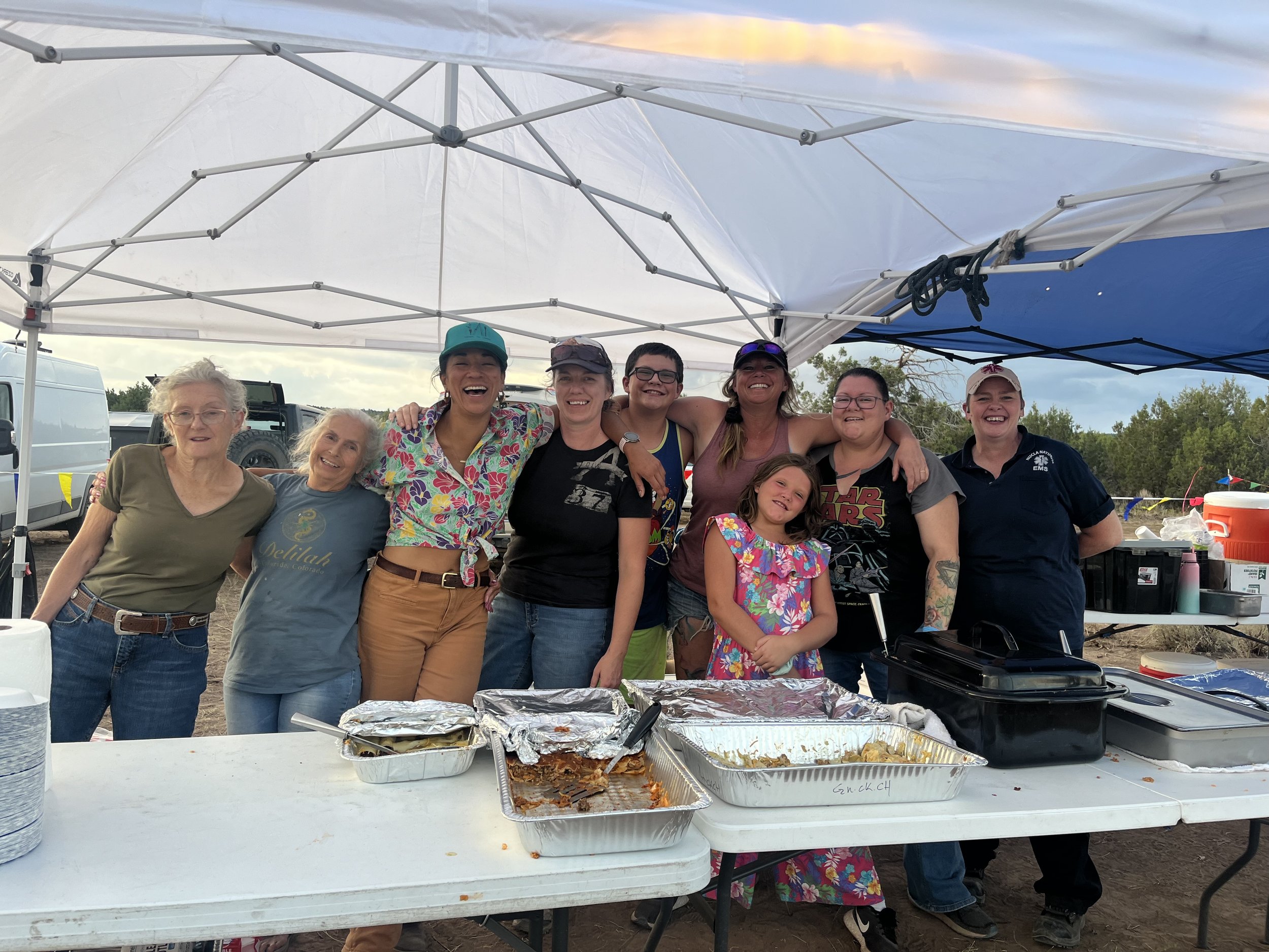 Un grupo de nueve personas sonríe bajo un toldo blanco, con bandejas de comida sobre una mesa. Llevan ropa informal y hay una niña entre ellos. El escenario parece ser una reunión social al aire libre.