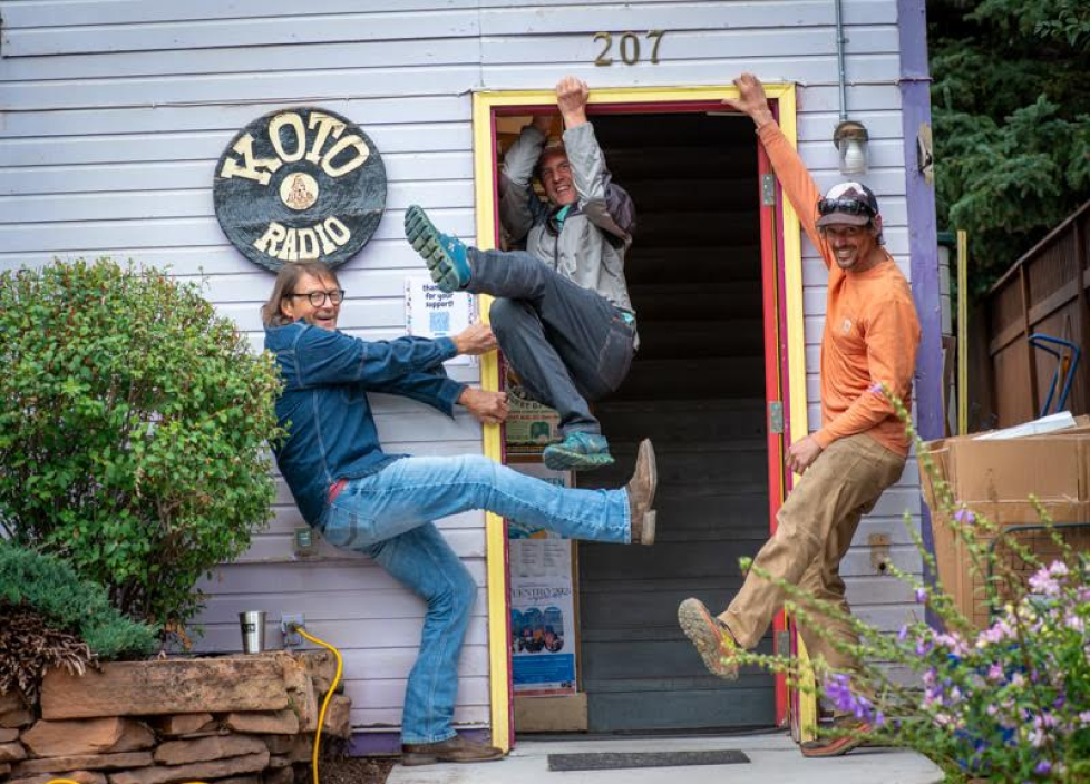 Three people joyfully performing high kicks at the entrance of KOTO Radio, with one person holding the door open. They are casually dressed and smiling, surrounded by a small garden and wooden wall with the stations sign.