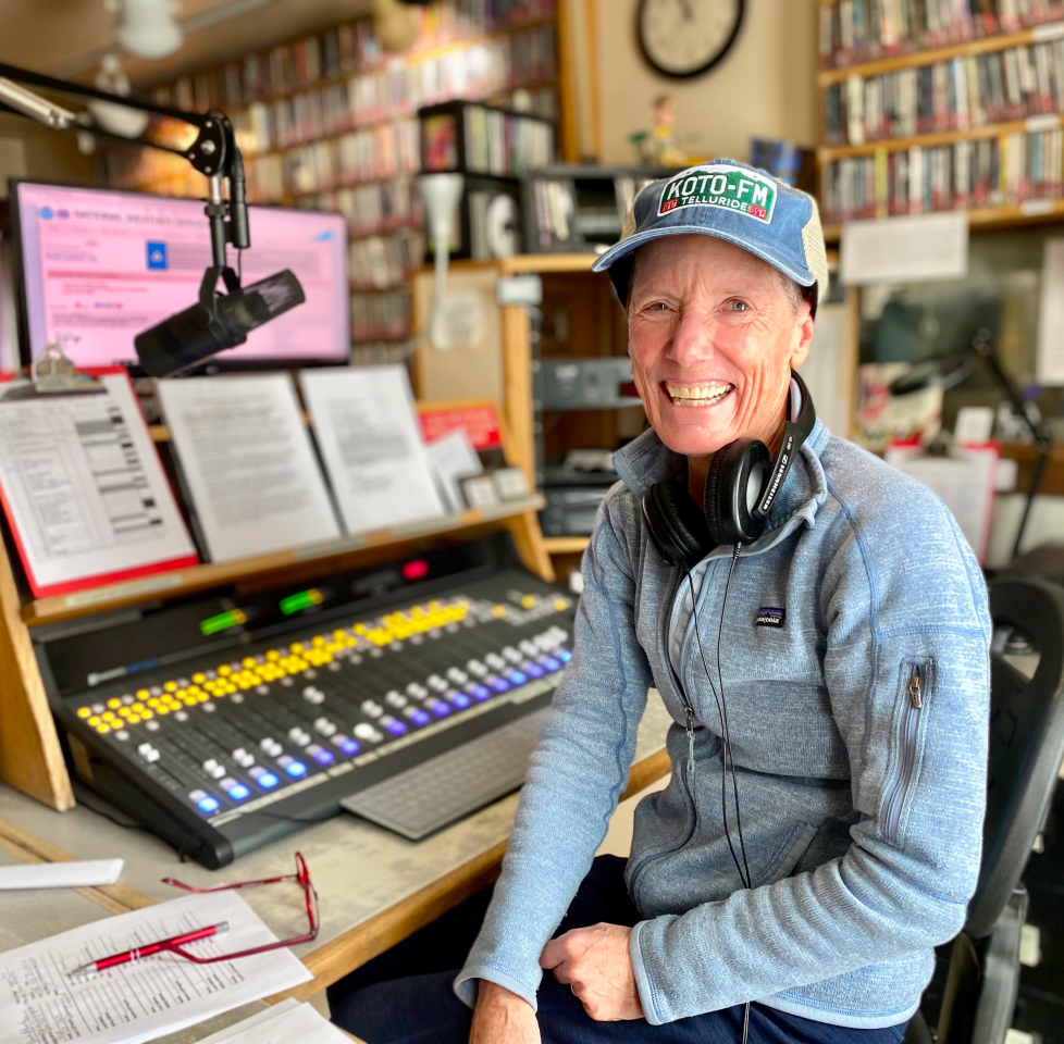 Person smiling in a radio station studio, wearing a blue cap and gray sweater. Headphones are around their neck. A microphone, papers, and colorful soundboard are visible. Shelves with CDs fill the background.