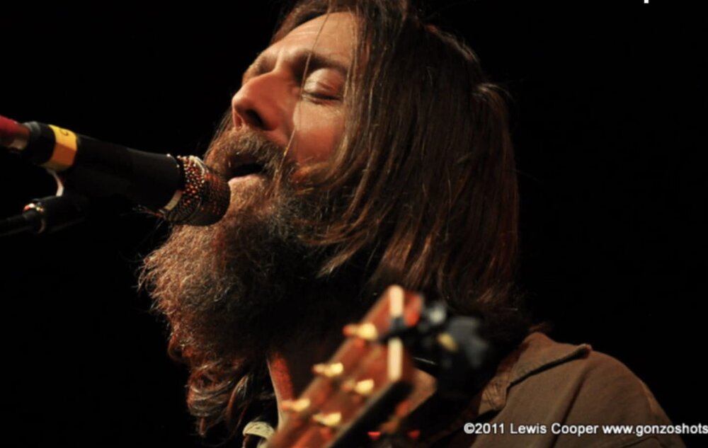 A bearded man with long hair sings into a microphone on stage, eyes closed, holding a guitar. The image is captioned with ©2011 Lewis Cooper www.gonzoshots.com.