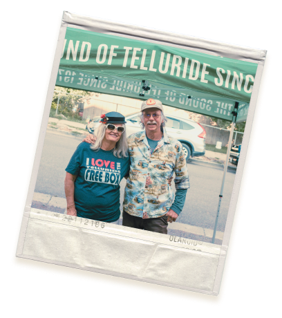 Two people stand under a tent with Telluride visible. The person on the left wears sunglasses, a dark hat, and a shirt with I love my free box. The person on the right wears a light floral shirt and a cap. Both smile at the camera.