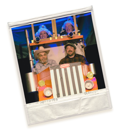 A Polaroid image of a playful setup with four people behind a fake truck front. Two people are visible through the windshield wearing wigs and hats, and two in the front seat wearing cowboy hats, all appearing to be having fun.