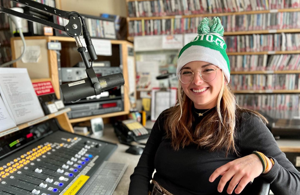 Young woman wearing glasses and a green knit hat, smiling in a radio studio. She is seated next to audio equipment with numerous CDs on shelves in the background.