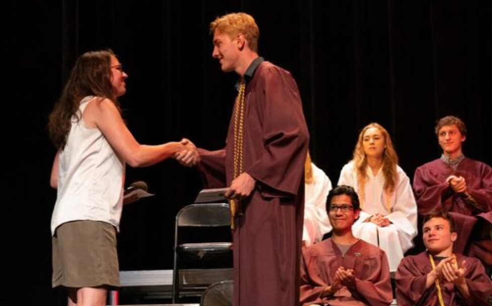 A graduate in a maroon robe shakes hands with a teacher on stage during a ceremony. Other graduates in maroon and white robes sit and clap in the background.