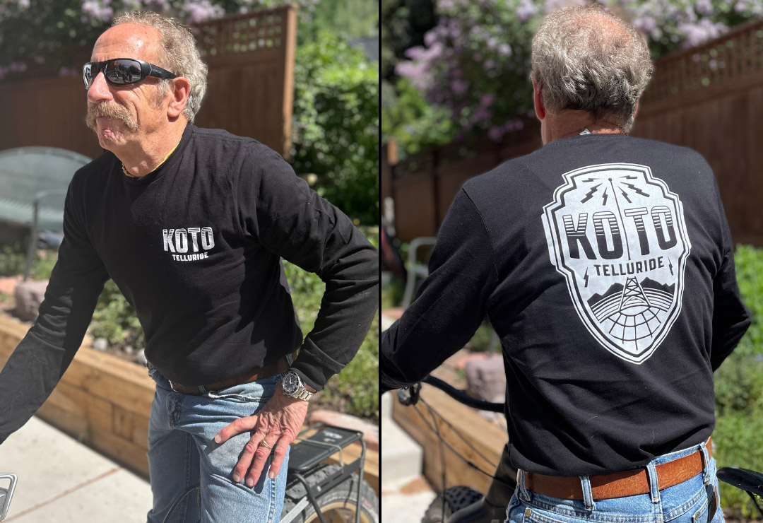 A man with a mustache wears sunglasses and a black KOTO Telluride shirt. He is seen from the front and back, standing near a bicycle with a wooden fence and trees in the background.