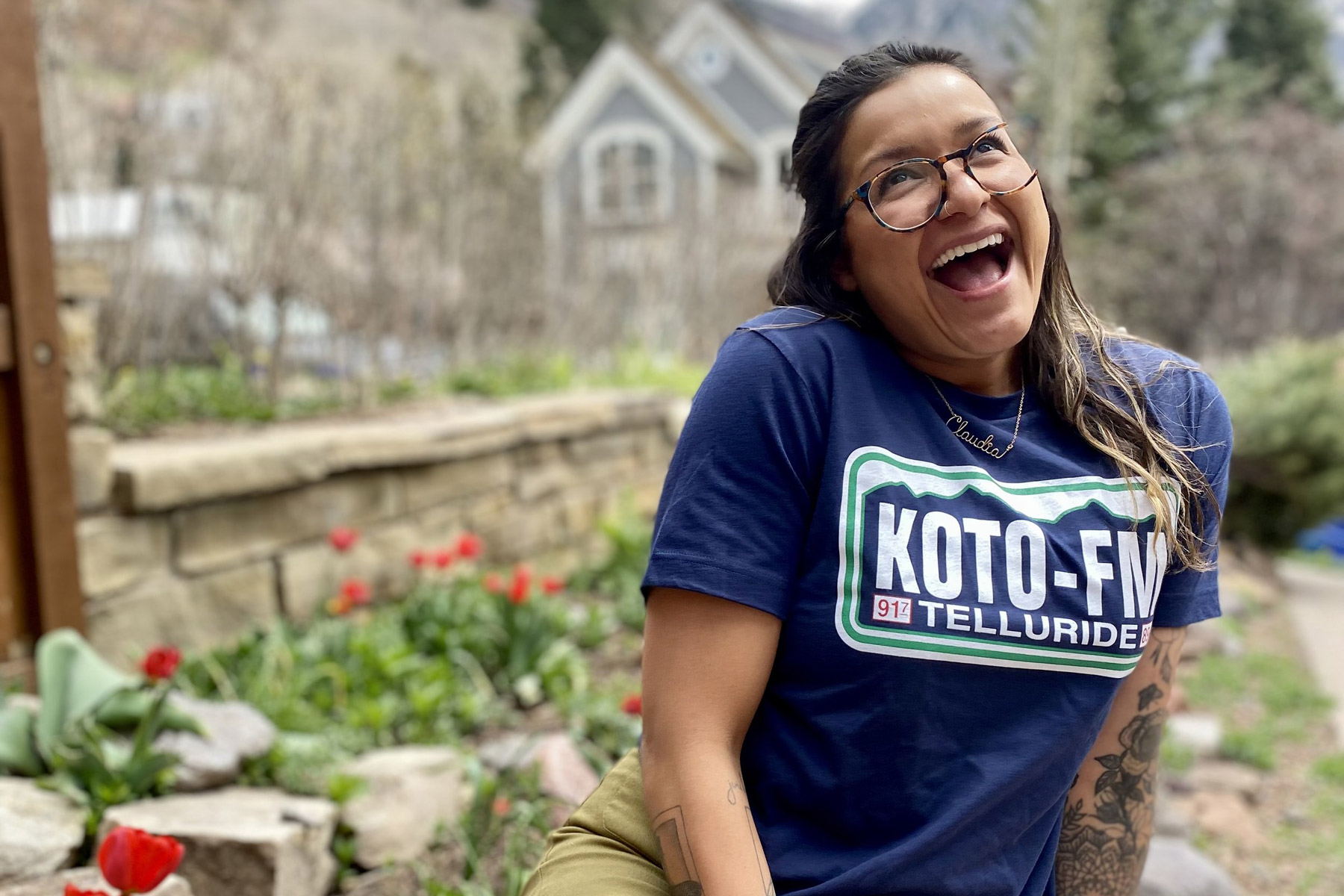 A woman with glasses smiles broadly while sitting outdoors. She is wearing a blue KOTO-FM Telluride shirt. Behind her are red tulips and a stone wall, with a blurred background of greenery and a house.