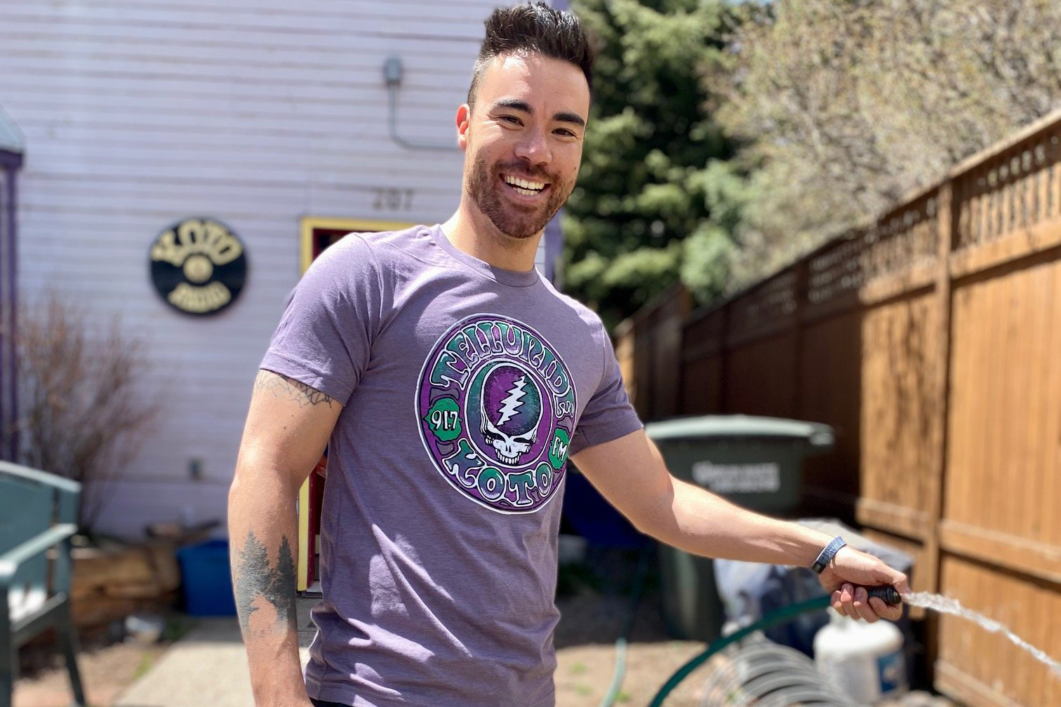 A person with short hair and a beard smiles while holding a hose in a sunny backyard. They are wearing a purple T-shirt with a graphic design. A fence, a few trees, and a building with a circular sign are in the background.