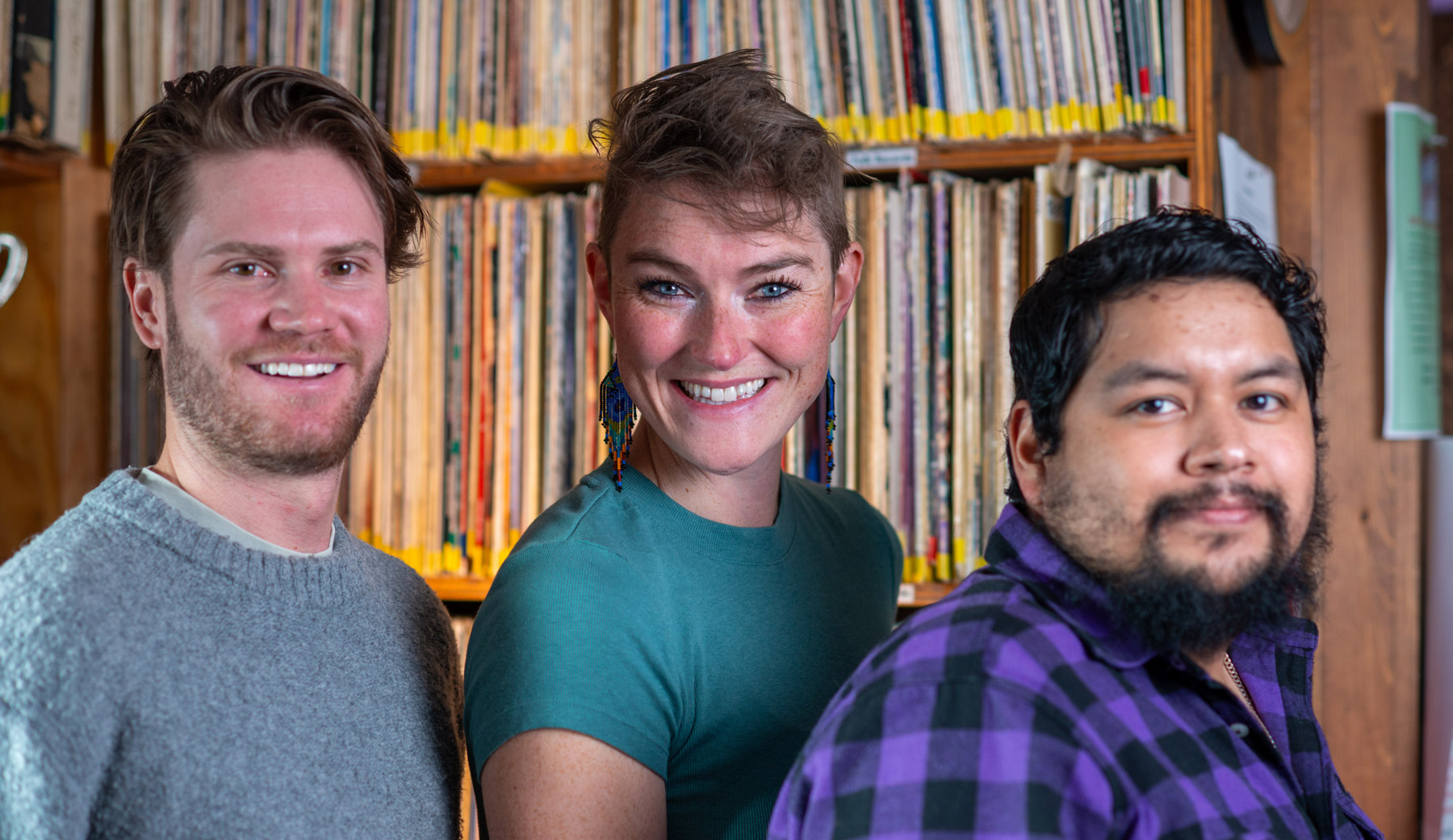 KOTO News team standing in front of shelves filled with records. They are smiling at the camera. The person on the left is wearing a gray sweater, the one in the middle is in a teal shirt, and the person on the right is wearing a purple plaid shirt.