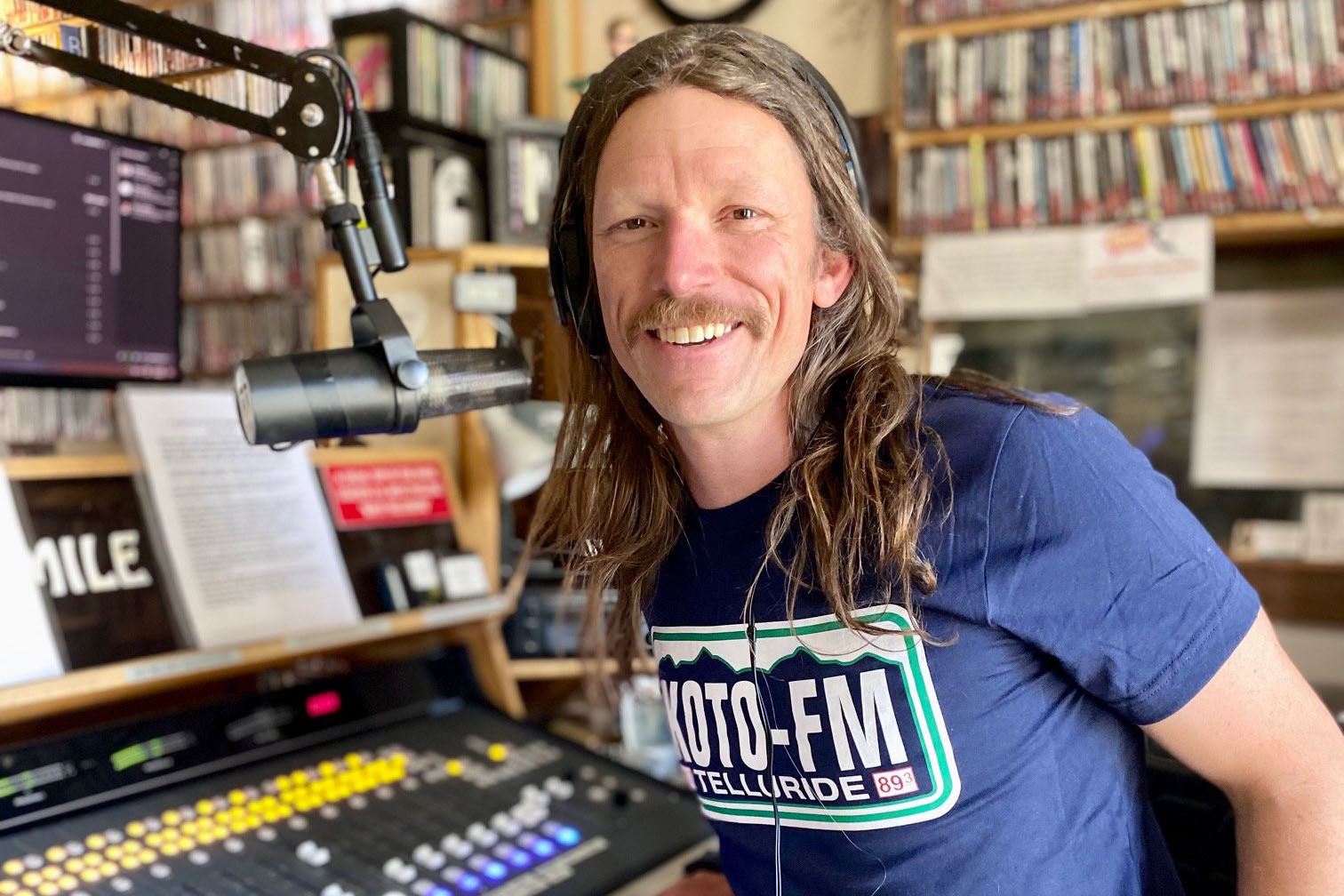 A person with long hair and a mustache is smiling while wearing headphones in a radio station. They are standing in front of a microphone and equipment, surrounded by shelves filled with CDs. The persons shirt reads KOTO-FM Telluride.