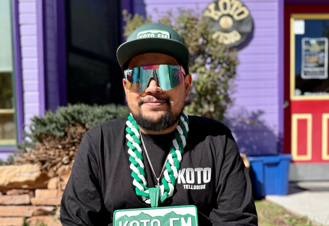 A person wearing reflective sunglasses, a green cap and black shirt with KOTO lettering, stands outside a purple building. They have a large green and white chain necklace, holding a green KOTO FM sign.