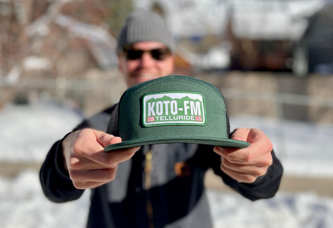 Person outdoors holding a green hat with a KOTO-FM Telluride logo. The background is snowy, and the person is wearing a gray beanie and sunglasses. The image is focused on the hat.