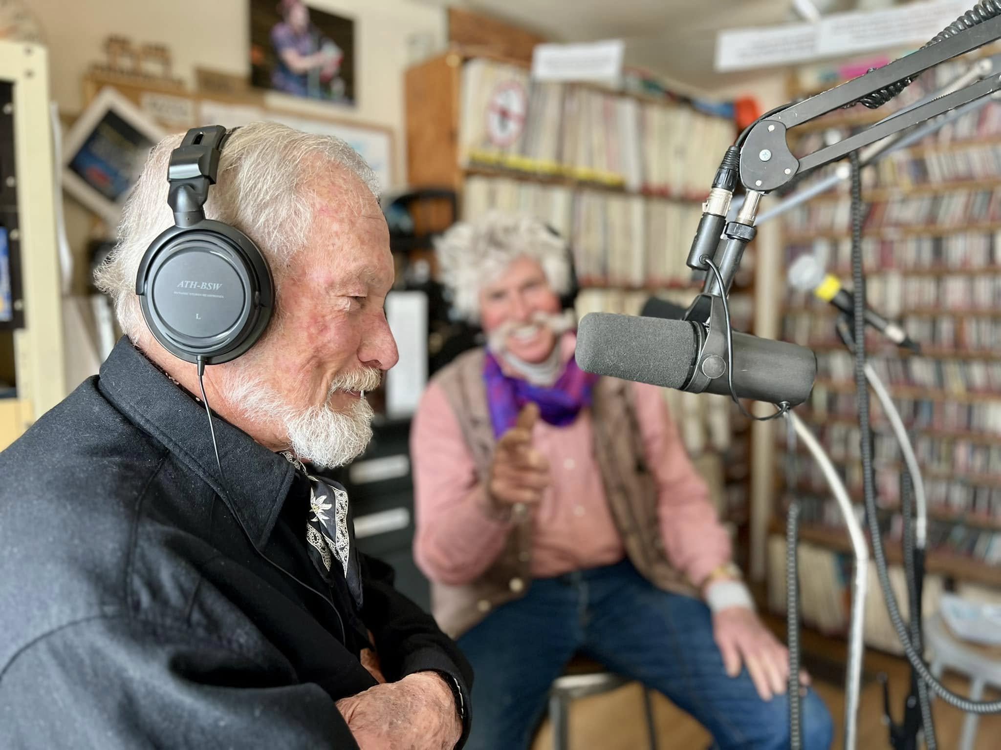 Two men in a radio studio. The man in the foreground wears headphones, a black jacket, and has crossed arms while speaking into a microphone. The man in the background, with a playful expression, points towards him. Shelves of records are visible behind them.