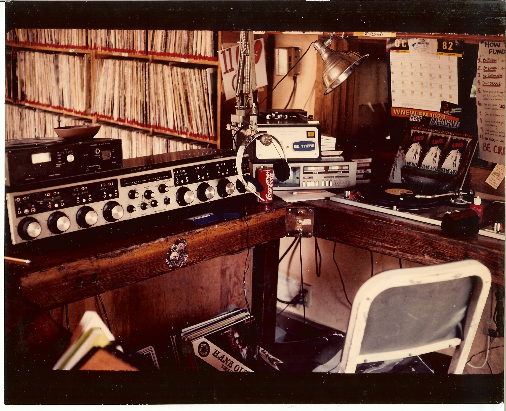 A vintage radio station studio with a large sound mixer, record player, tape deck, and equipment on a wooden desk. Shelves of vinyl records line the background. A microphone and headphones are set up, with a chair in front of the desk.