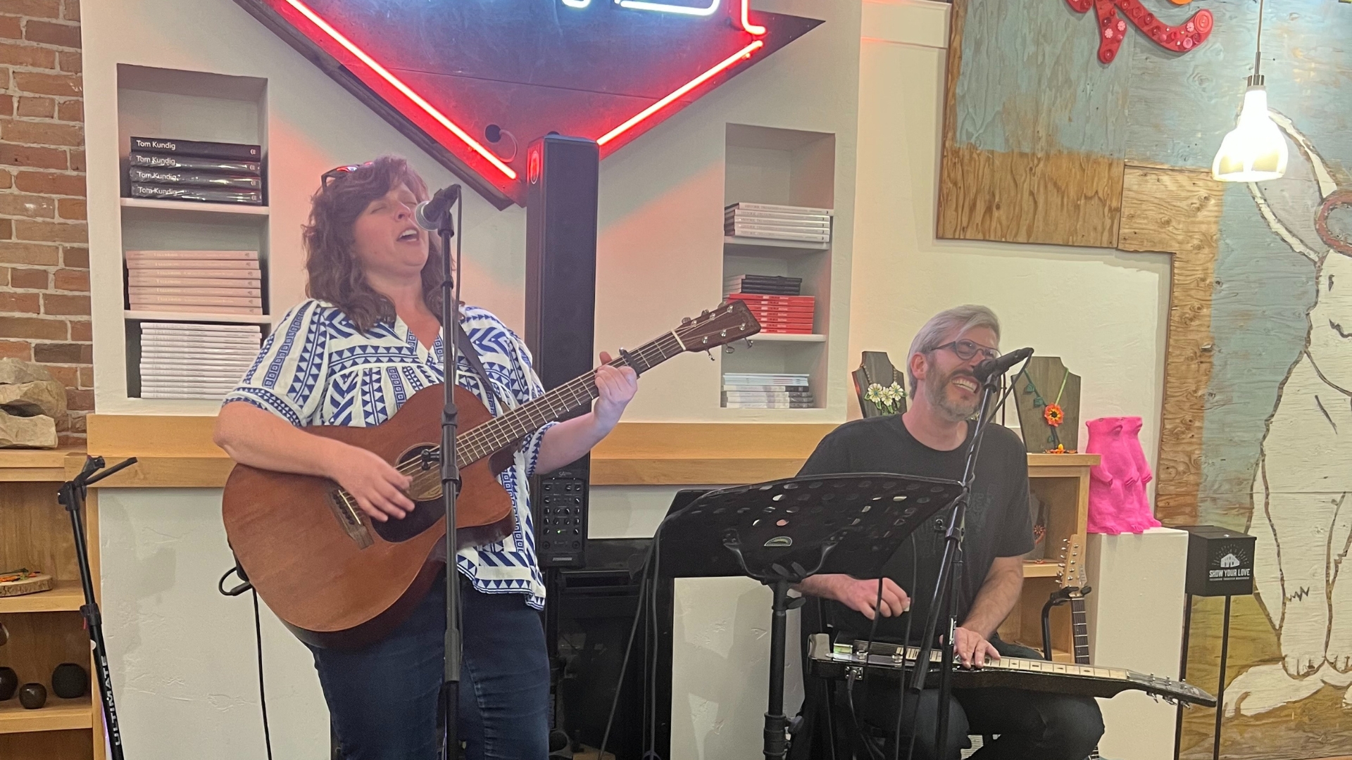 Two musicians perform; a woman sings and plays guitar, while a man, seated, plays an instrument with a slide. Theyre in a cozy room with brick and wood walls, a red neon sign, and various decorative items around. Both appear joyful and engaged in the music.