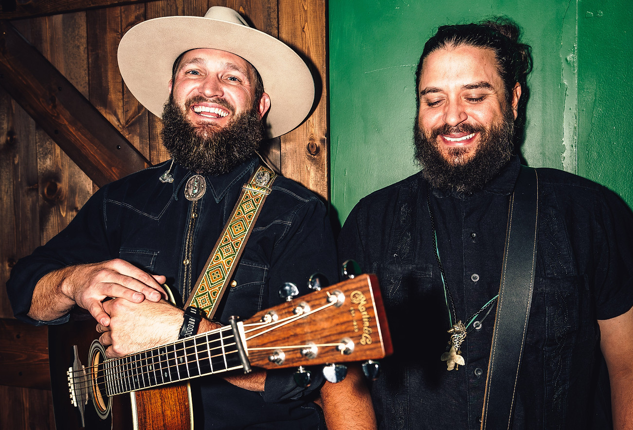 Two men with beards are standing and smiling. The man on the left is wearing a wide-brimmed hat and holding a guitar, while the man on the right is wearing a long necklace. They are in a room with wooden and green walls.