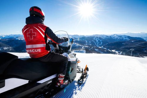 Una persona con un chaleco rojo de rescate conduce una moto de nieve por una montaña nevada e iluminada por el sol. El fondo muestra un vasto paisaje de picos nevados y un cielo azul claro y brillante.