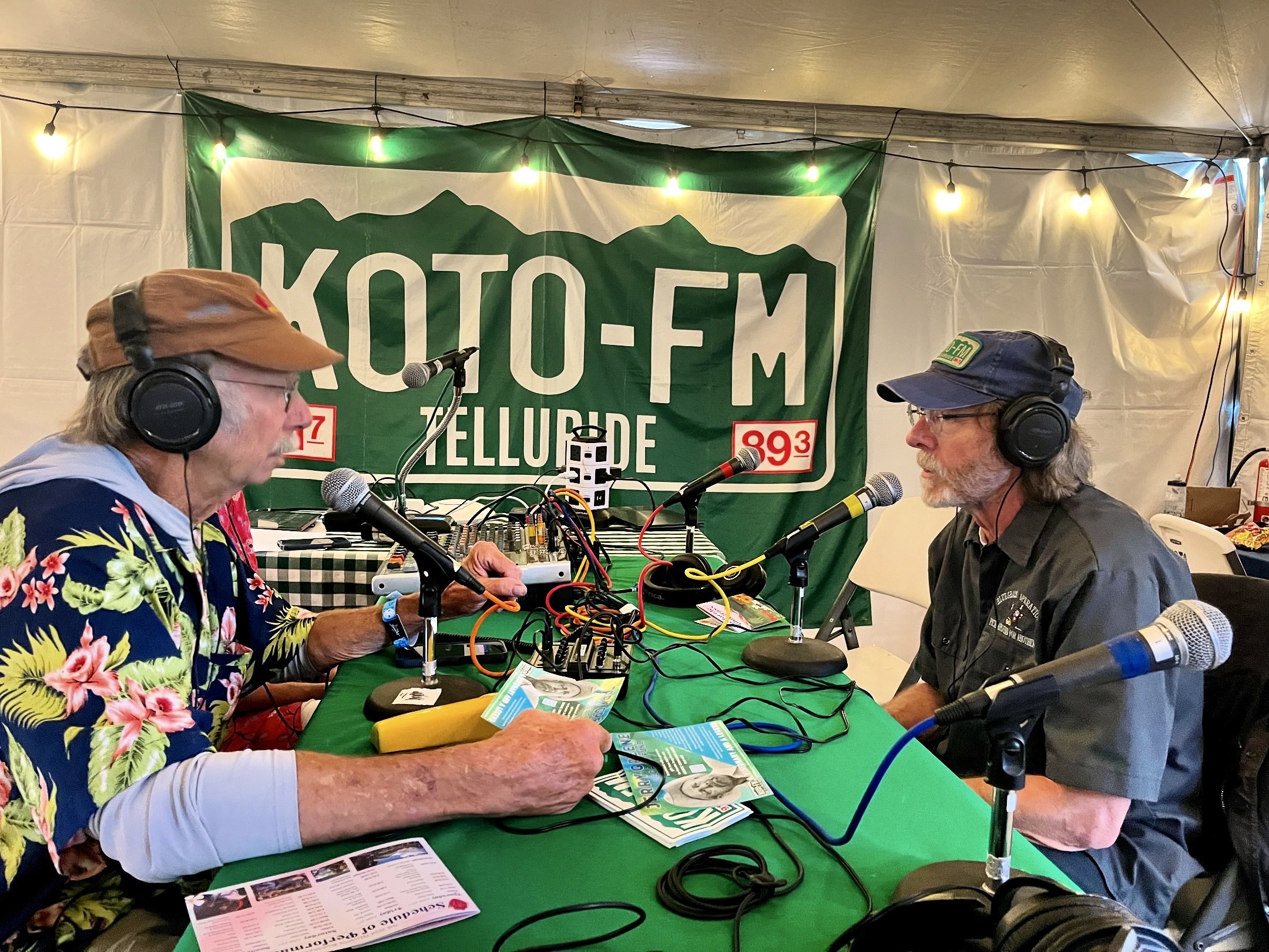 Two men sit across from each other speaking into microphones at a radio setup. Both wear hats, with one in a floral shirt and the other in a black shirt. A banner for KOTO-FM Telluride hangs behind them, with cables and papers on the table.