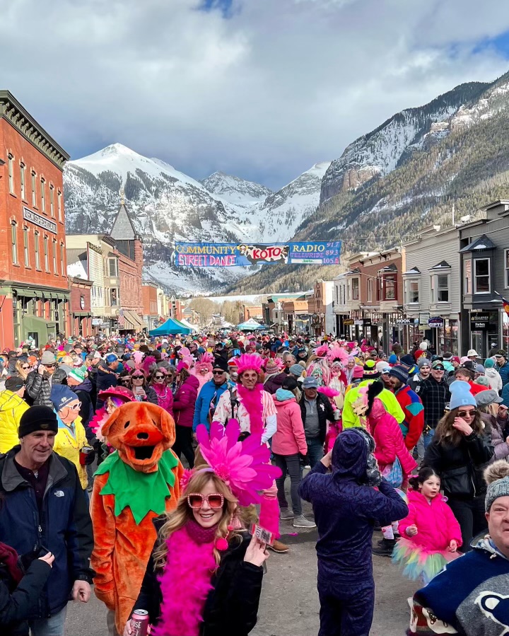 A lively street festival in a mountain town, with people in colorful costumes, including pink feathers and an orange animal suit. Snow-capped peaks and buildings line the street, and a banner hangs overhead. The sky is partly cloudy.