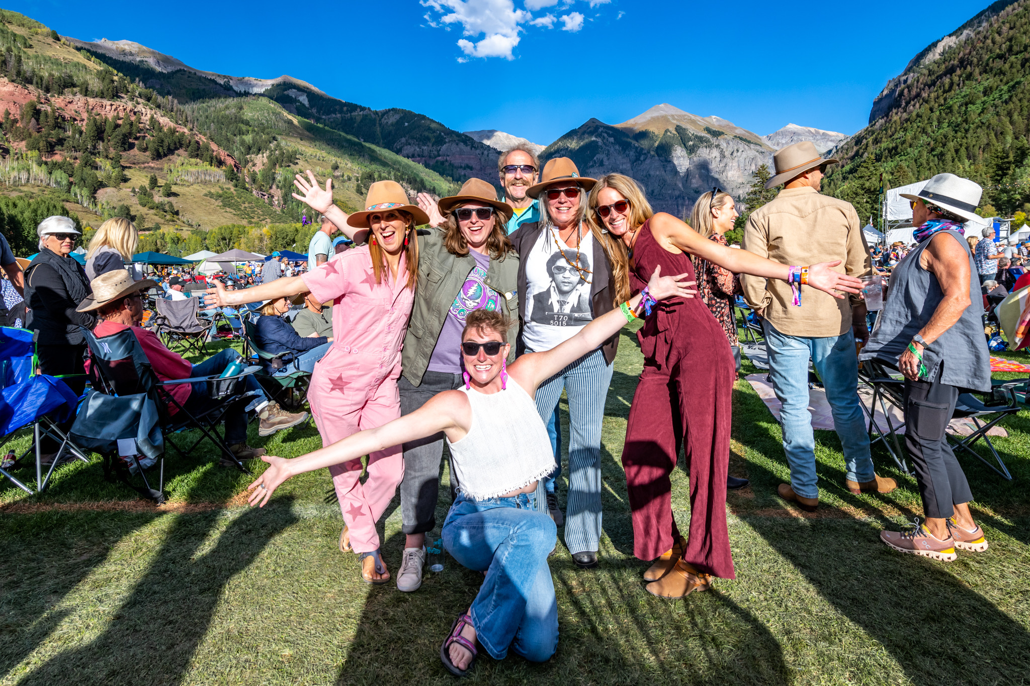 A group of six people posing cheerfully at an outdoor festival. They are dressed casually in summer attire, with mountains and a clear blue sky in the background. Some are wearing hats and sunglasses, and there are people seated on the grass nearby.