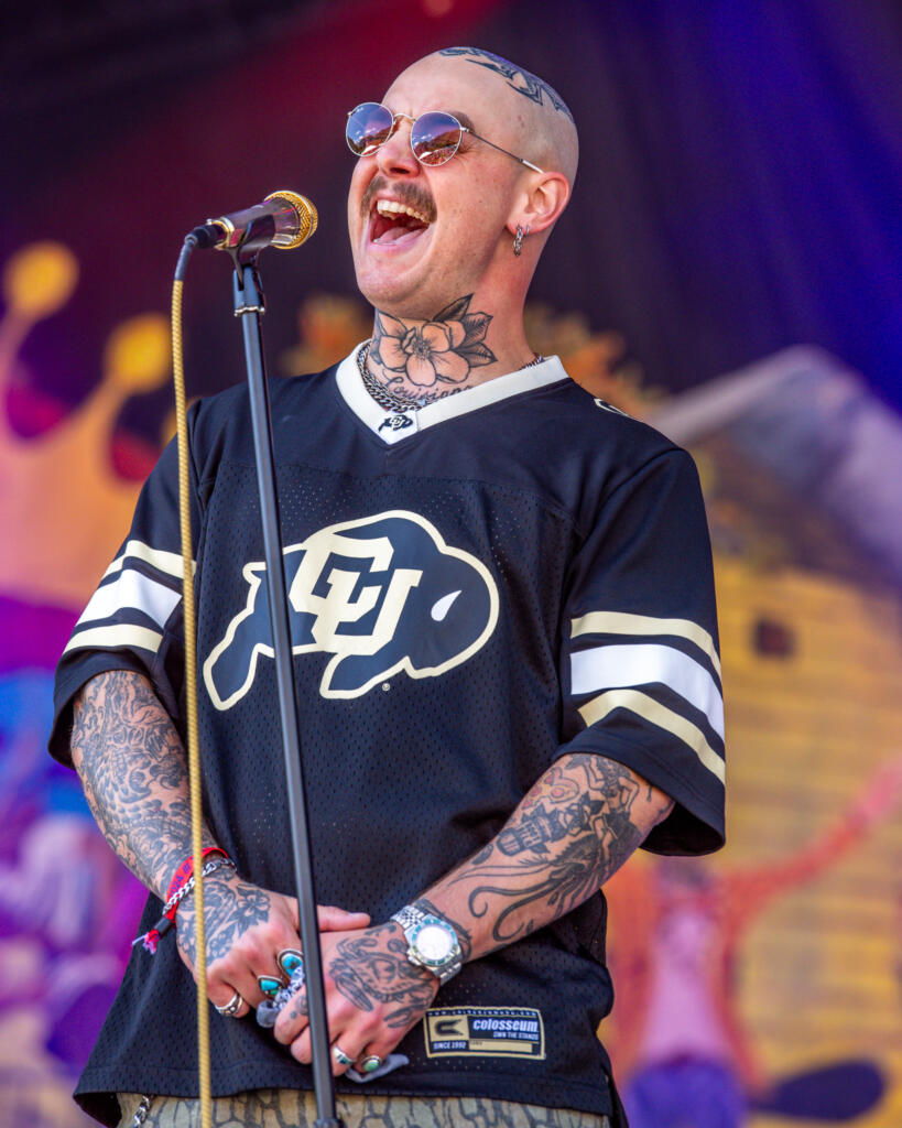 A person with sunglasses and tattoos is singing passionately into a microphone. They are wearing a black Colorado Buffaloes jersey and have a mustache. The background is colorful and out of focus.