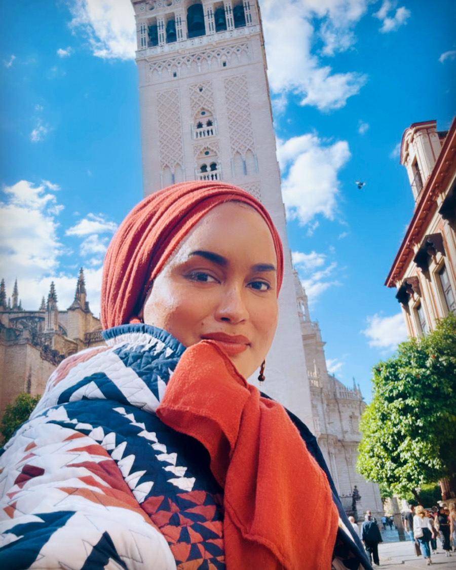 A person wearing a red headscarf and a colorful jacket stands smiling in front of a tall, ornate tower against a blue sky with clouds. The architecture suggests a historic or cultural site. Trees and buildings surround the area.