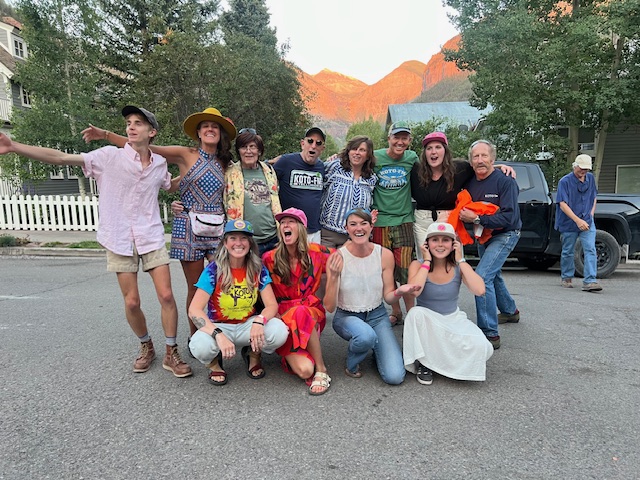 A group of people posing happily outdoors on a street. They are dressed in casual, colorful attire, with some wearing hats. In the background, there are trees, houses, and mountains under a clear sky.