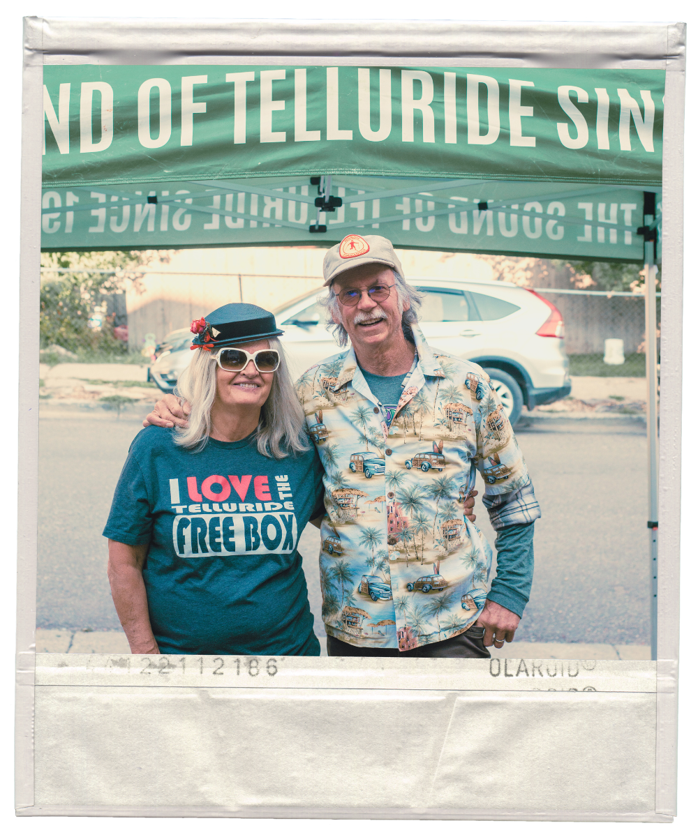 Dos personas sonríen bajo una carpa verde. La persona de la izquierda lleva gafas de sol, una gorra negra y una camiseta azul I Love the Free Box. La persona de la derecha lleva gafas, gorra y una camiseta estampada. El fondo muestra un coche y parte de un cartel.