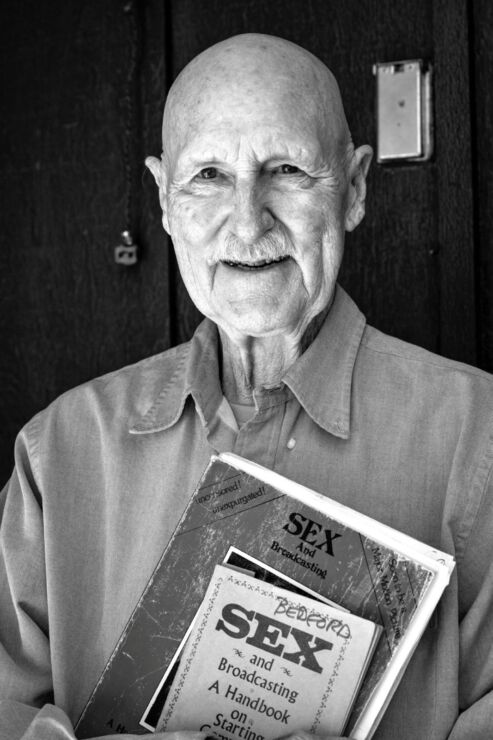 20250325-Z73_0084-Edit An elderly man with a mustache, co-founder of KOTO-FM, holds a stack of books and papers—including one titled Sex and Broadcasting—standing in front of a dark wooden door.