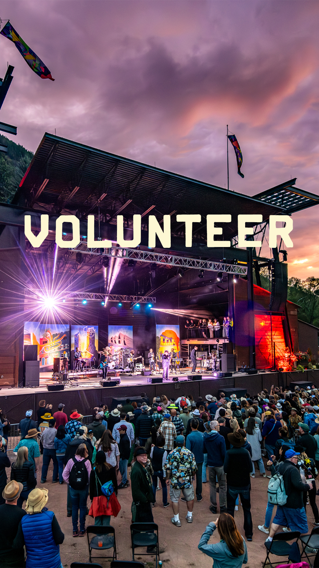 A crowd watches a live outdoor concert at sunset during the Telluride Blues & Brews Festival, with the word VOLUNTEER displayed in large letters above the stage.