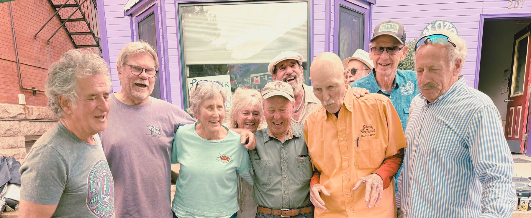 A group of older adults standing together and smiling outside a purple building with large windows and a red door.