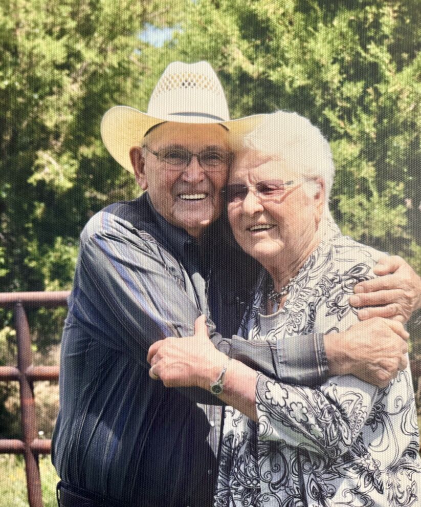 An elderly man in a cowboy hat, identified as Kirk Alexander, hugs an elderly woman outdoors. Both are smiling at the camera with trees in the background.