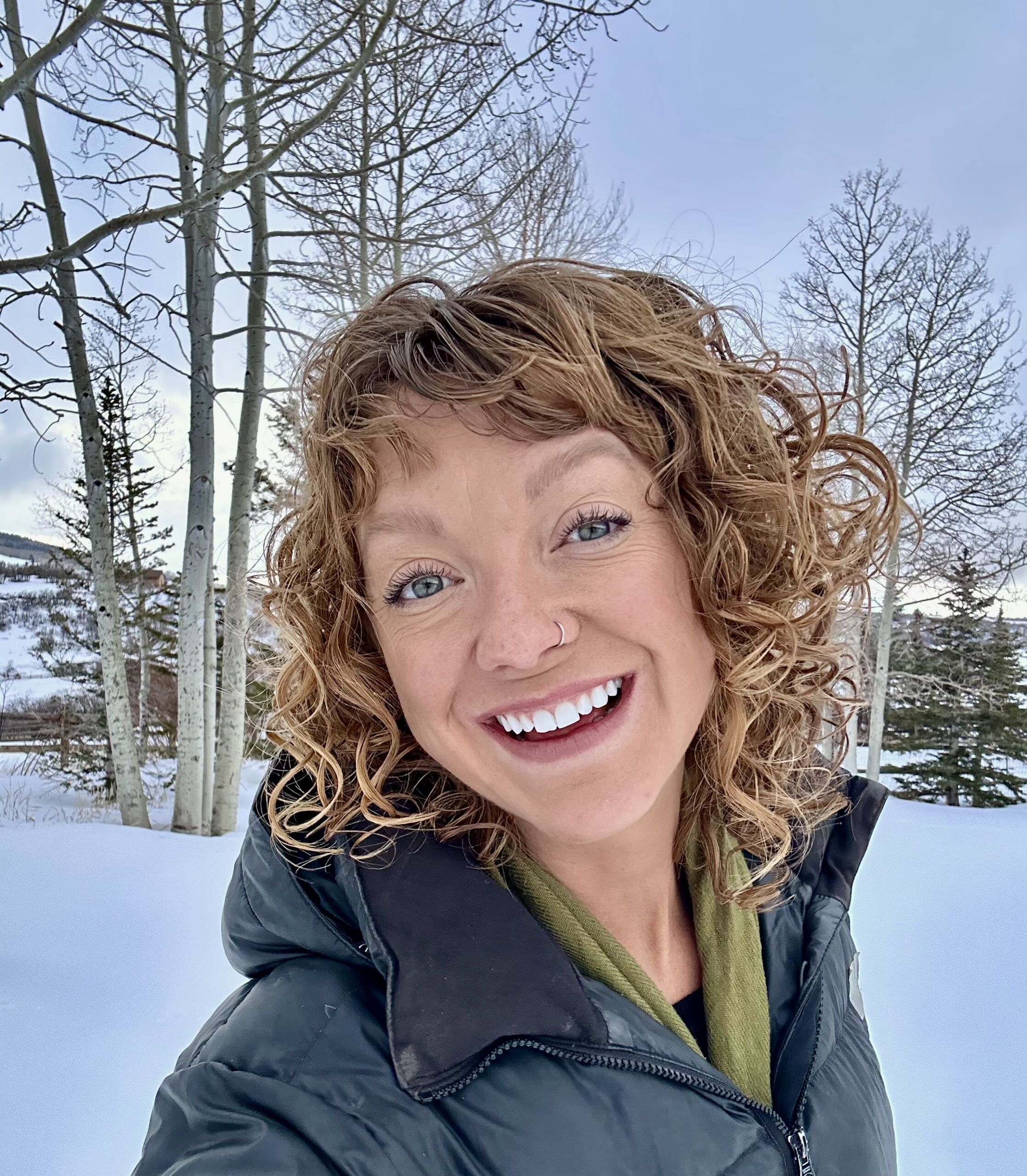 A person with curly hair and a nose ring smiles outdoors amid frosty snow, with trees in the background, embracing the solstice chill.