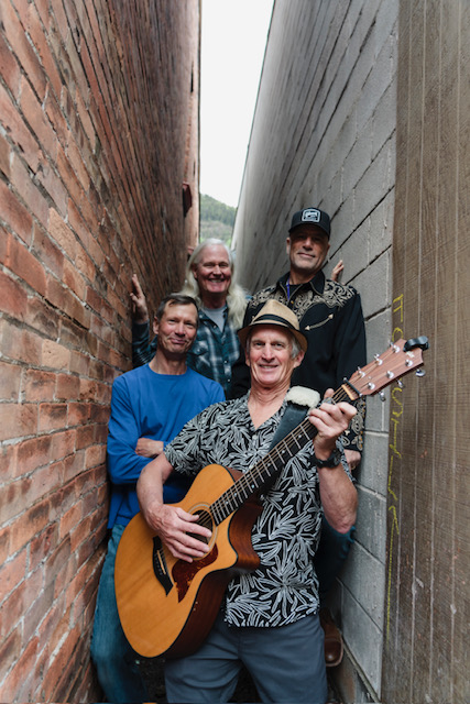 Four men stand closely together in a narrow alleyway, with the frontman of the Telluride Gold Kings holding an acoustic guitar and smiling at the camera—ready to bring Saint Patrick's Day cheer.