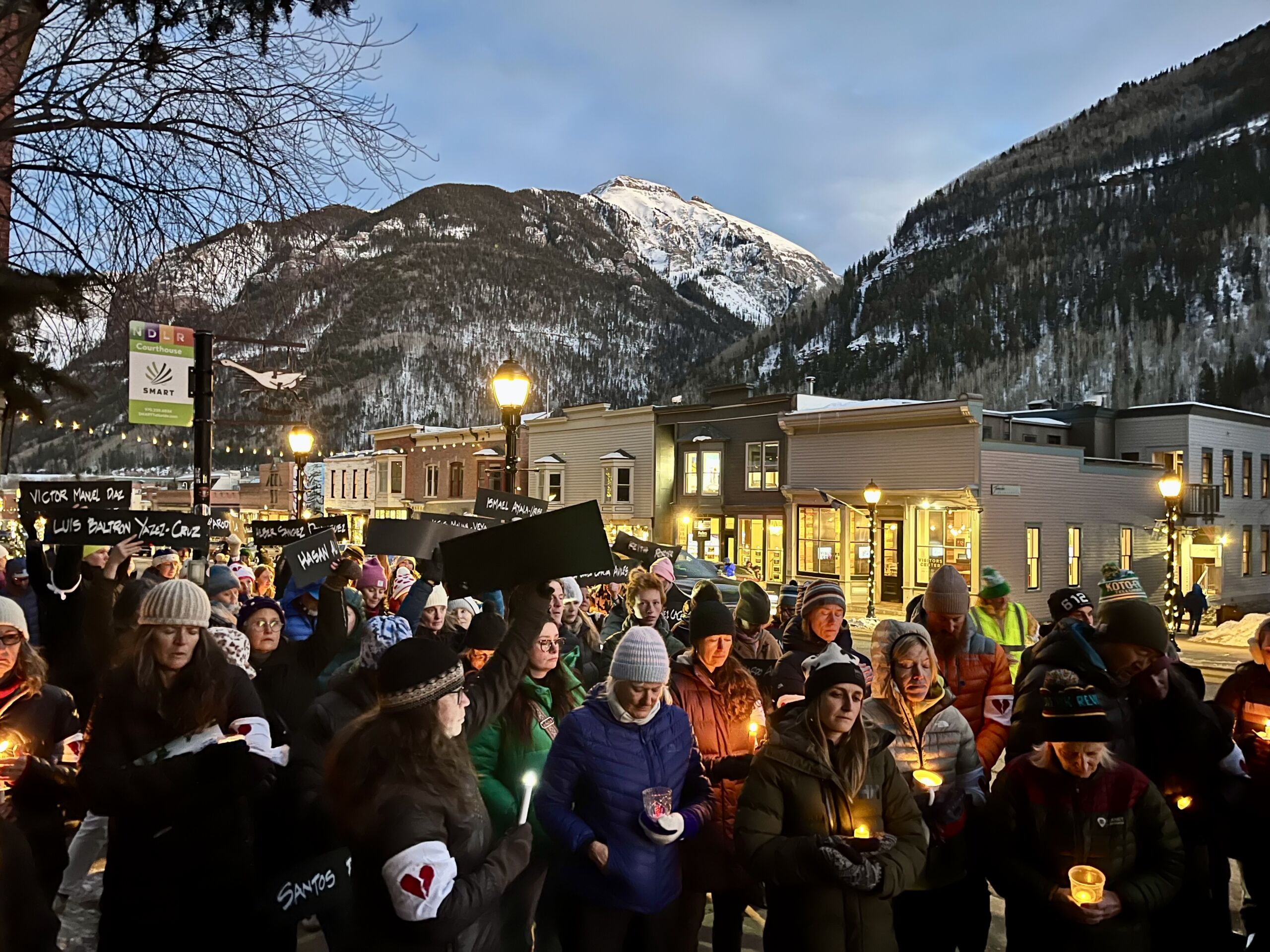 A group of people holding candles gathers outdoors in a snowy mountain town at dusk, participating in a candlelight vigil that honors loved ones amidst the serene beauty of ice and snow.