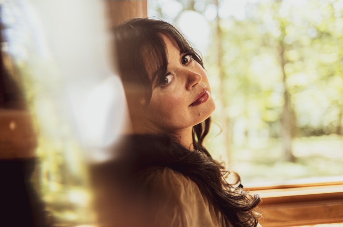 A woman with long dark hair sits indoors by a window, looking over her shoulder toward the camera, a sense of intimacy and wonder in her gaze, with sunlight and Appalachia’s lush trees visible outside.