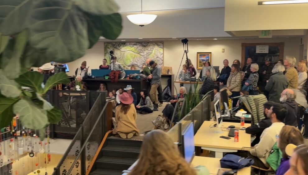 A group gathered inside Wilkinson Public Library, some seated on stairs and others standing, enjoying a Tiny Circ Desk Concert near a piano; computer desks are visible in the foreground.