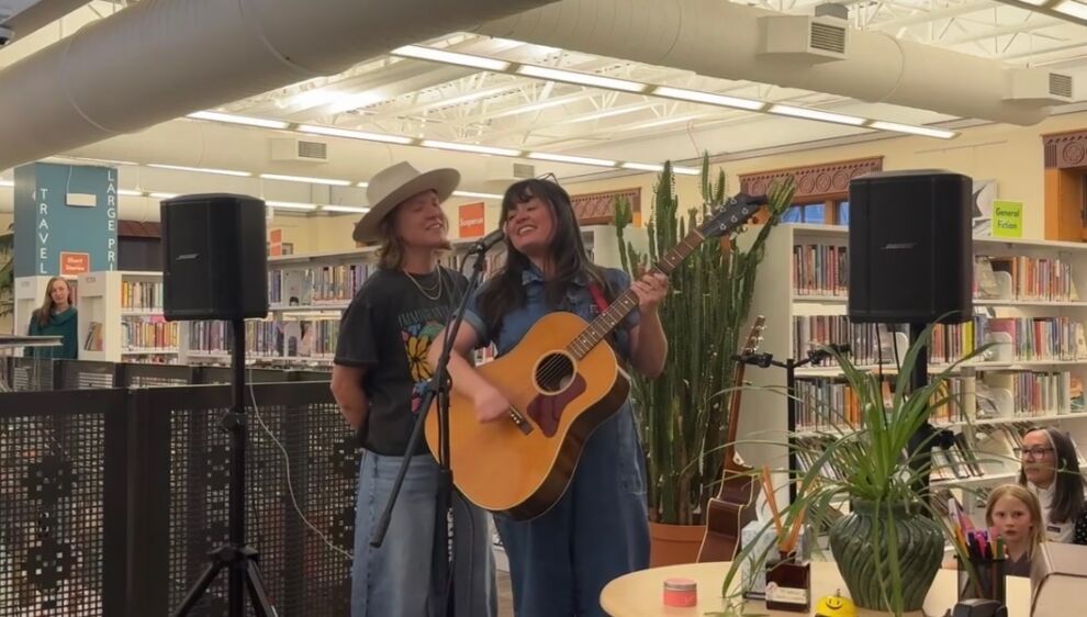 Two people perform music at Wilkinson Public Library’s Tiny Circ Desk Concerts; one plays guitar and sings while the other stands smiling. Bookshelves and plants serve as a charming backdrop.