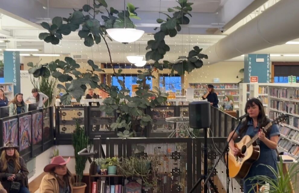 A woman plays an acoustic guitar and sings into a microphone at Wilkinson Public Library, surrounded by bookshelves, plants, and an audience, as part of the intimate Tiny Circ Desk Concerts series.